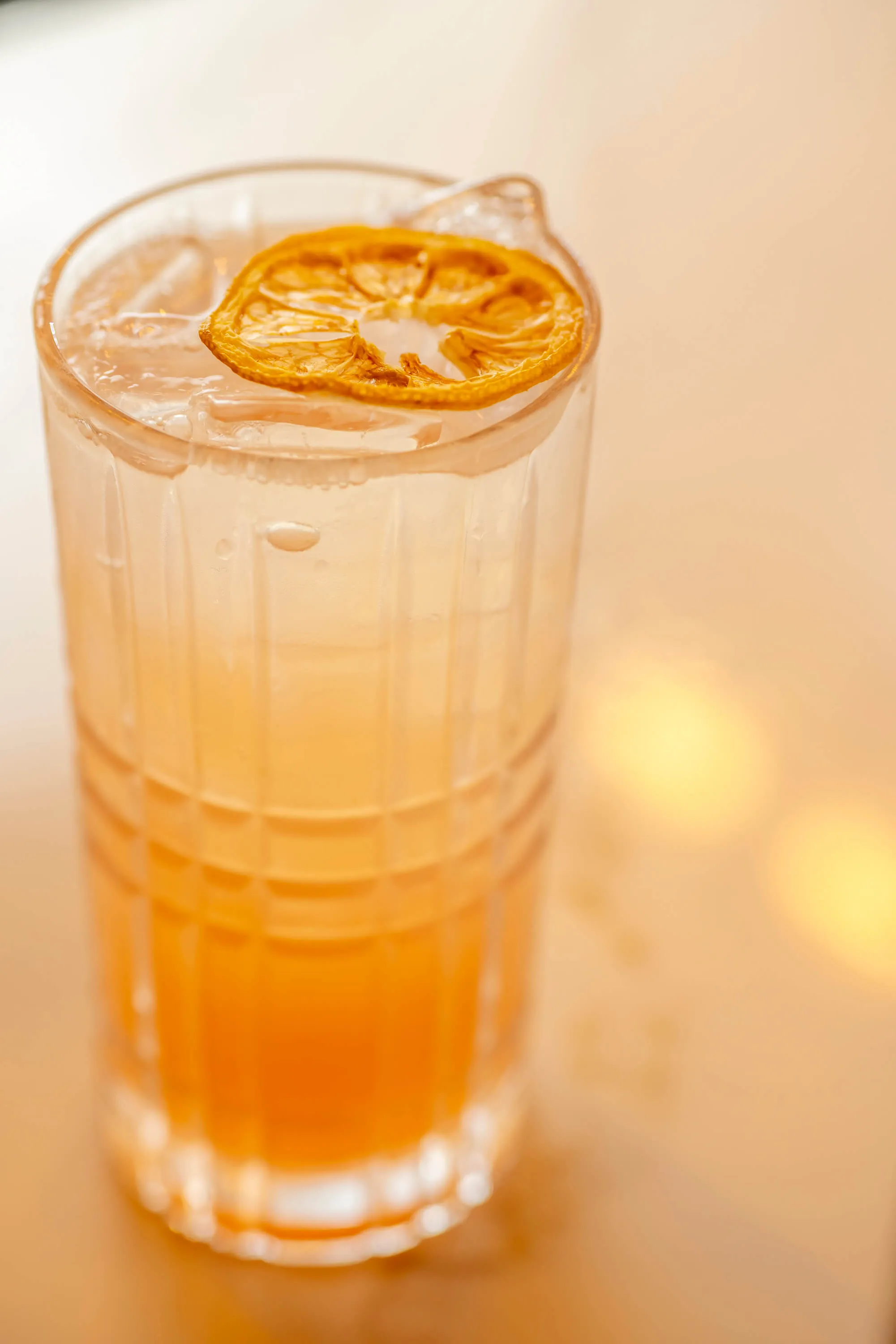 Tall glass with an orange gradient drink, ice cubes, and a dried orange slice garnish on top.